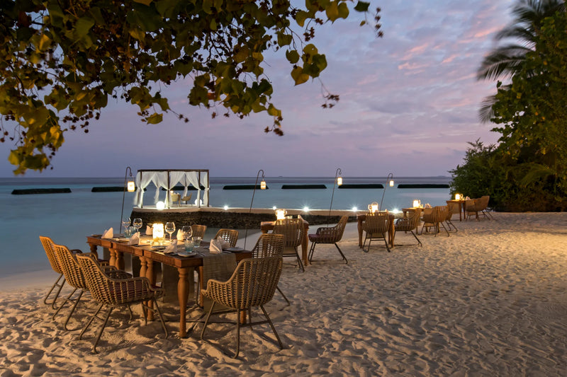 Constance Moofushi, South Ari Atoll: dinner tables set on the sand at dusk, candlelit, facing the lagoon
