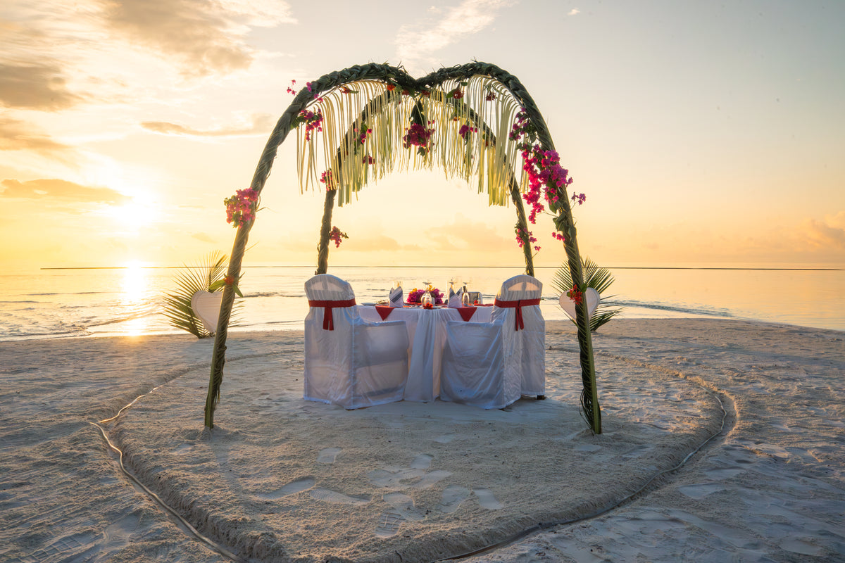 A dinner table for two under a flower arch on the beach at sunset, Medhufushi Island Resort.