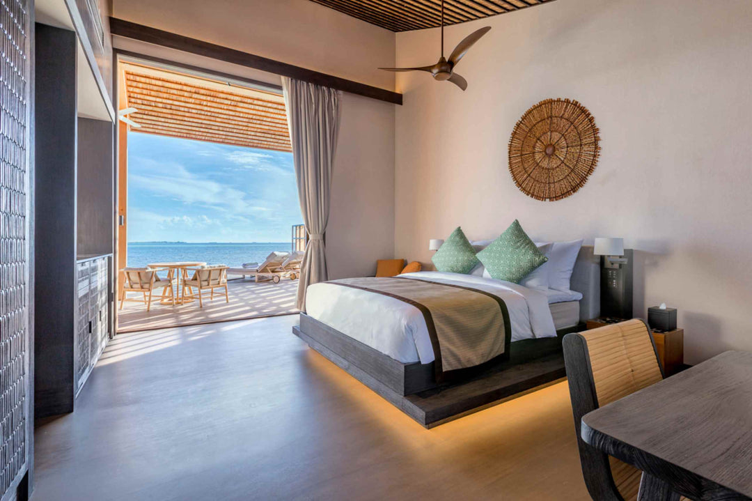Kudadoo Maldives Private Island, Lhaviyani Atoll — residence bedroom looking out through open sliding doors to the deck, dining table and loungers, with the lagoon beyond