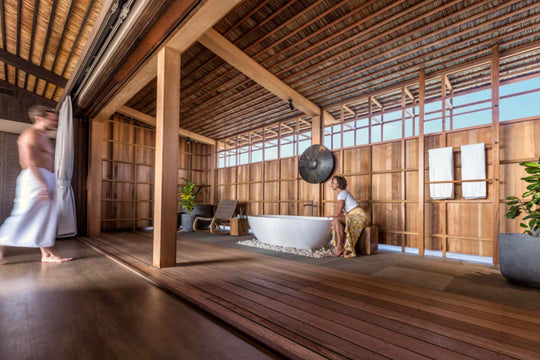 Kudadoo Maldives Private Island, Lhaviyani Atoll — open-sided bathroom with a freestanding bath set on pebbles, timber-slatted screens and guests nearby