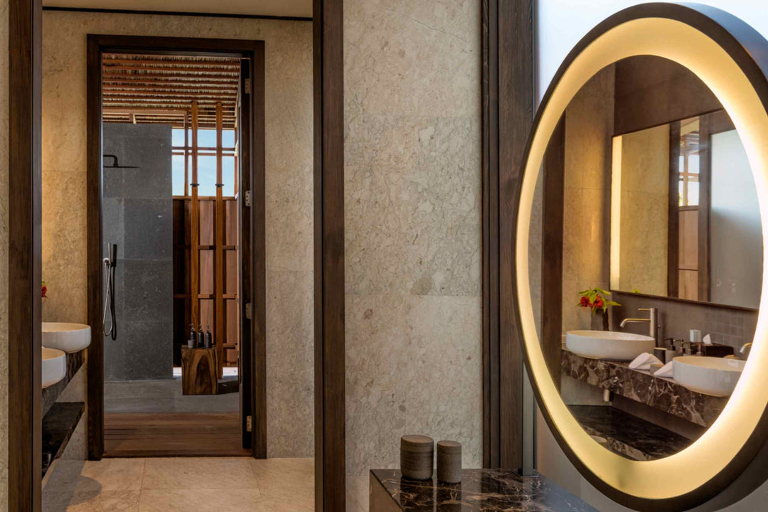 Kudadoo Maldives Private Island, Lhaviyani Atoll — bathroom with twin stone basins, a backlit oval mirror and a walk-in rain shower through the doorway
