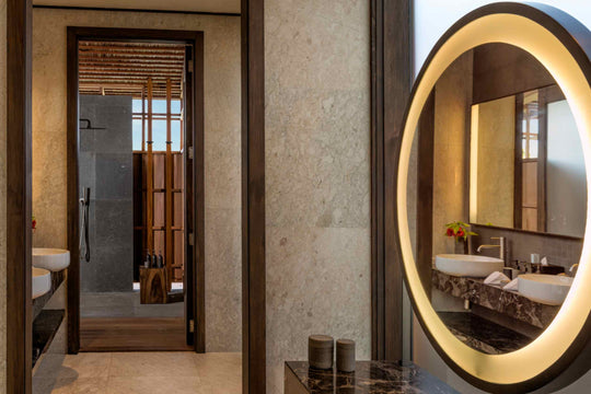 Kudadoo Maldives Private Island, Lhaviyani Atoll — bathroom with twin stone basins, a backlit oval mirror and a walk-in rain shower through the doorway