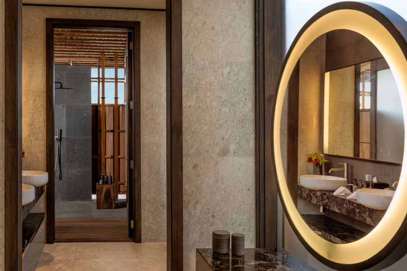A bathroom at Kudadoo Maldives Private Island with twin stone basins, a backlit oval mirror and a walk-in rain shower through the doorway.