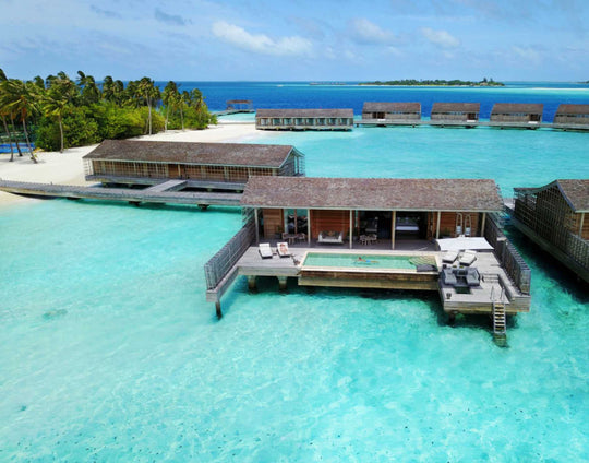 Kudadoo Maldives Private Island, Lhaviyani Atoll — aerial view of overwater residences and the main building along the lagoon edge, with the beach and palms behind