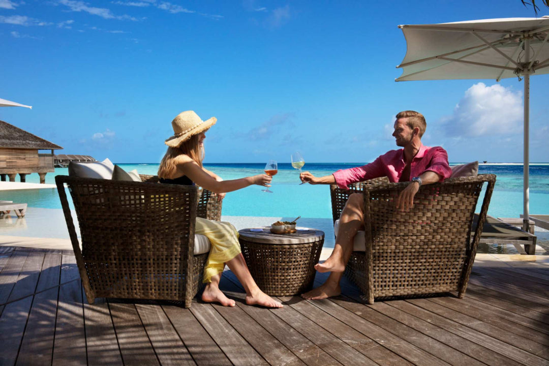 Kudadoo Maldives Private Island, Lhaviyani Atoll — a couple in wicker armchairs on the deck raising glasses of wine, with the pool and ocean behind them