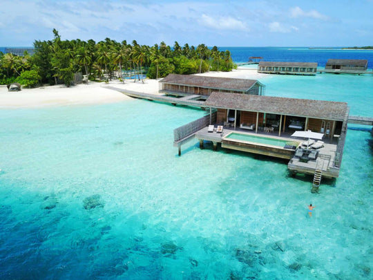 Kudadoo Maldives Private Island, Lhaviyani Atoll — aerial view of the palm-covered island, its white beach and the overwater residences reaching across the lagoon
