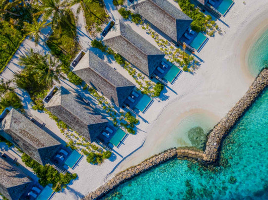 Kagi Maldives Resort &amp; Spa, North Malé Atoll — aerial view of a row of beach villas with shingled roofs and private pools, set behind the sand and a rock breakwater
