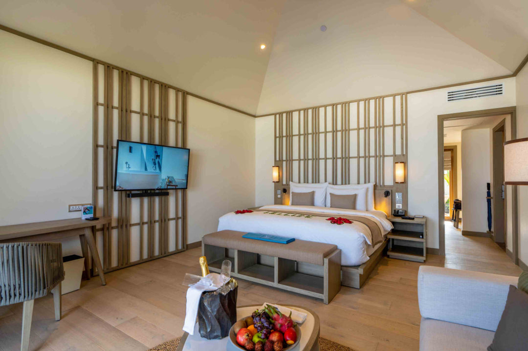 Kagi Maldives Resort &amp; Spa, North Malé Atoll — villa bedroom with a king bed dressed in red petals, timber slatted wall, wall-mounted television and a fruit platter and champagne on arrival