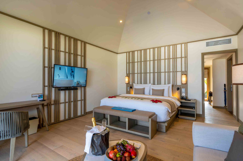 A villa bedroom at Kagi Maldives Resort & Spa with a king bed dressed in red petals, a timber slatted wall, a wall-mounted television and a fruit platter and champagne on arrival.