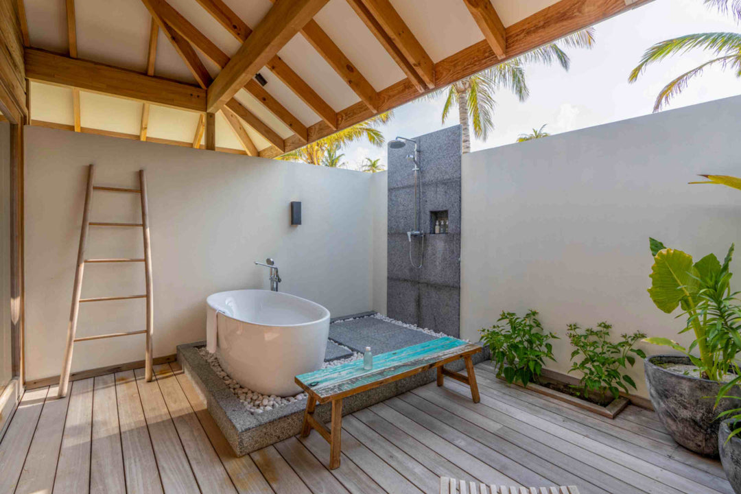 Kagi Maldives Resort &amp; Spa, North Malé Atoll — outdoor bathroom under a pitched timber roof, with a freestanding tub on pebbles, a stone rain shower and palms overhead