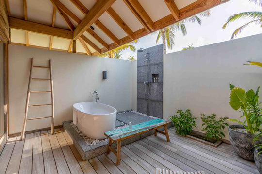 Kagi Maldives Resort &amp; Spa, North Malé Atoll — outdoor bathroom under a pitched timber roof, with a freestanding tub on pebbles, a stone rain shower and palms overhead