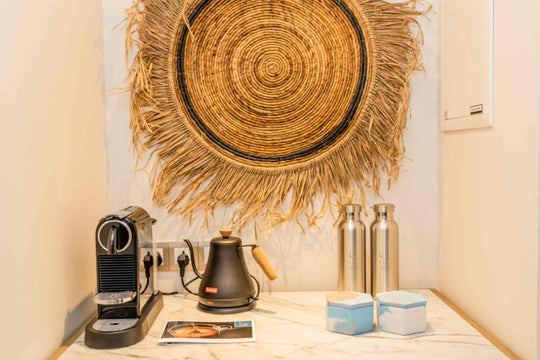 Kagi Maldives Resort &amp; Spa, North Malé Atoll — in-villa coffee station with a Nespresso machine, kettle and refillable water bottles beneath a woven palm wall disc
