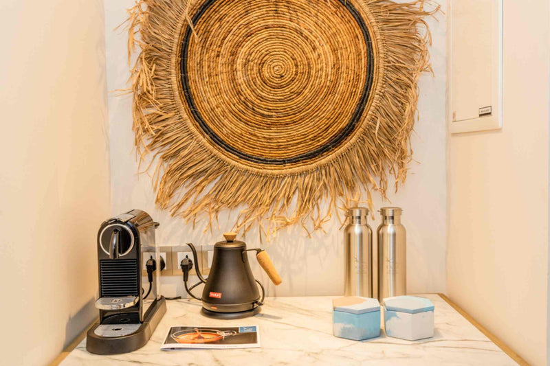 The in-villa coffee station at Kagi Maldives Resort & Spa, a Nespresso machine, kettle and refillable water bottles beneath a woven palm wall disc.