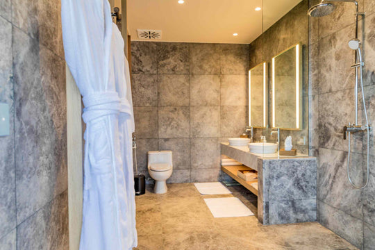 Kagi Maldives Resort &amp; Spa, North Malé Atoll — indoor bathroom in grey stone, with a robe hanging by the walk-in shower, twin basins and backlit mirrors