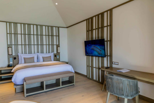 Kagi Maldives Resort &amp; Spa, North Malé Atoll — villa bedroom with a king bed against a slatted timber headboard wall, a bench at its foot and a writing desk