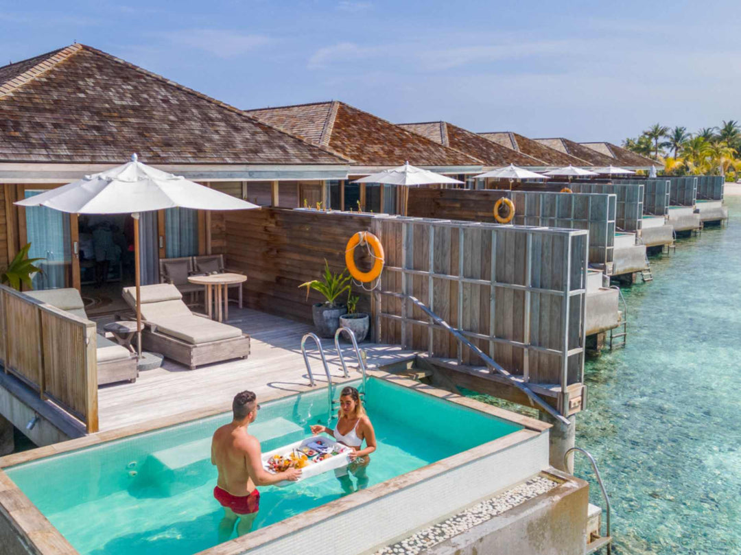 Kagi Maldives Resort &amp; Spa, North Malé Atoll — a couple sharing a floating breakfast tray in the private pool of an overwater villa, with the row of villas stretching behind