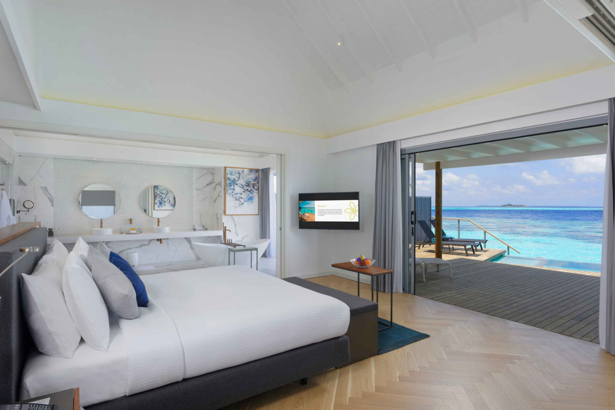 Over-water villa bedroom with king bed, open sliding doors and a deck facing the lagoon