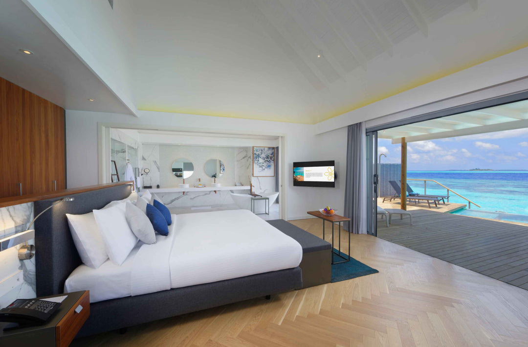 Over-water villa bedroom with a dark timber headboard and twin vanity visible behind the bed