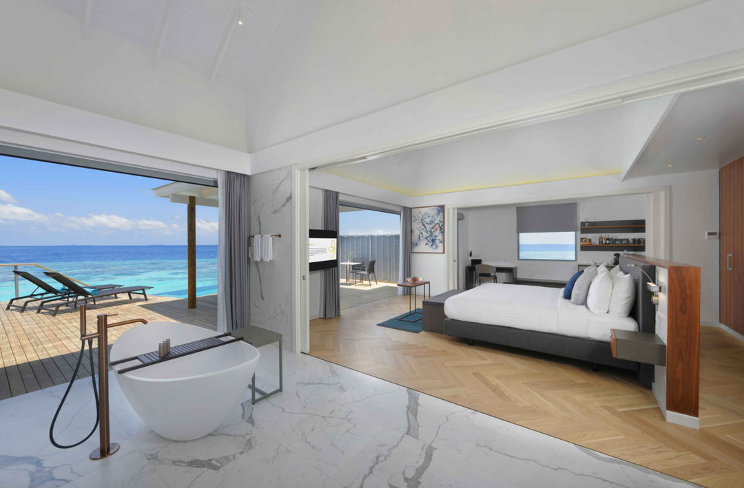 Over-water villa interior with freestanding bath, bed, and sun loungers on the deck beyond