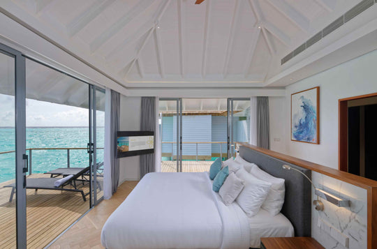 Over-water villa bedroom looking out through glass doors to the deck and lagoon