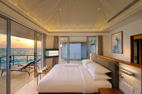 Over-water villa bedroom at sunset, the lagoon beyond the open glass doors