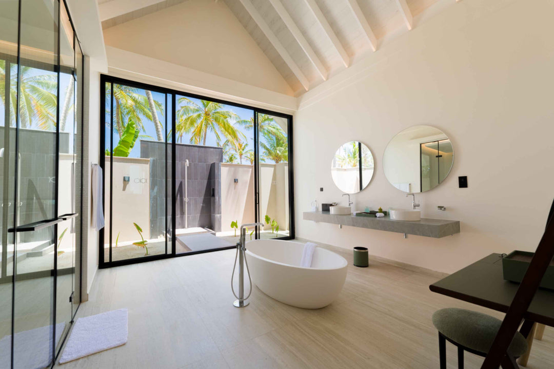 A villa bathroom at ananea Madivaru Maldives, bath and twin basins with glass doors to an outdoor shower