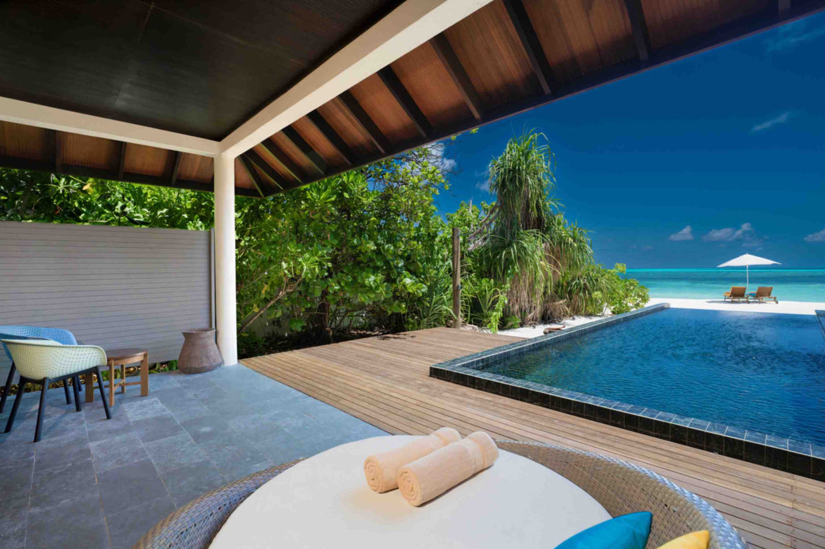 Atmosphere Kanifushi, Lhaviyani Atoll — Kanifushi Beach Villa with Pool: a shaded terrace with cane chairs beside a long private pool, then white sand, a parasol and the turquoise lagoon.