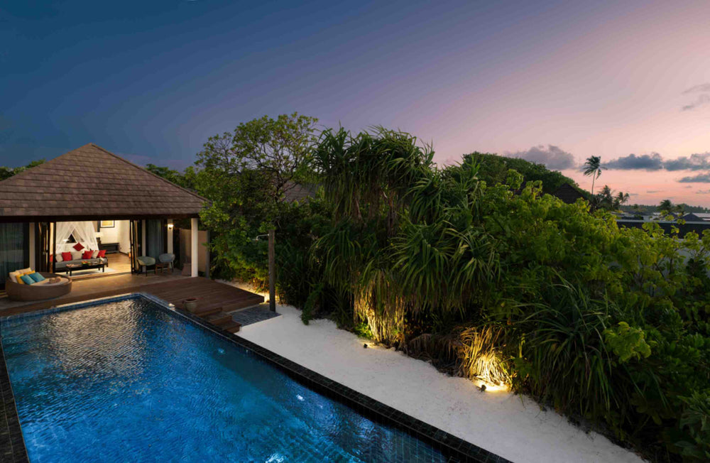 Atmosphere Kanifushi, Lhaviyani Atoll — Kanifushi Beach Villa with Pool at dusk, lit from inside behind its pool, screwpine and palms crowding the garden under a pink sky.