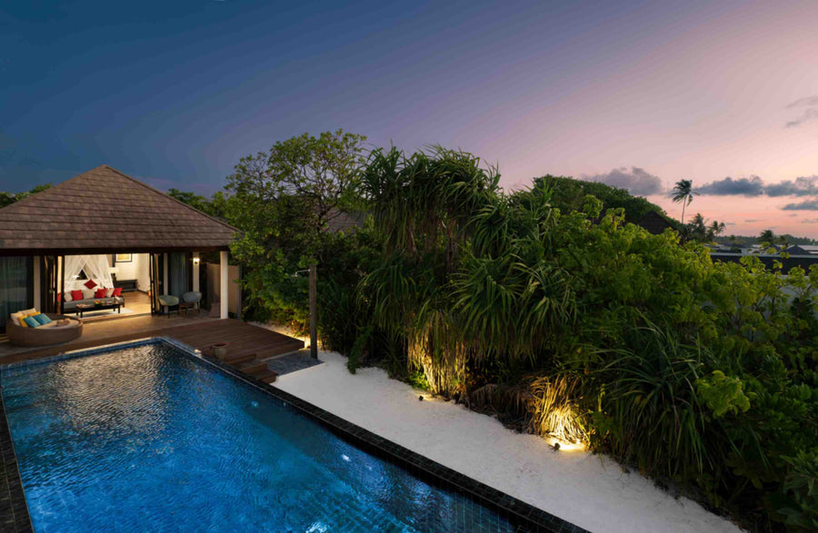 A Kanifushi Beach Villa with Pool at Atmosphere Kanifushi at dusk, lit from inside behind its pool, screwpine and palms crowding the garden under a pink sky.
