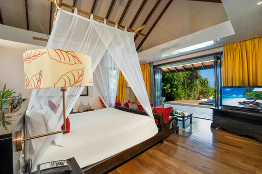 Atmosphere Kanifushi, Lhaviyani Atoll — a canopied bed in a Kanifushi Beach Villa with Pool, yellow curtains drawn back from glass doors on to the garden and pool.