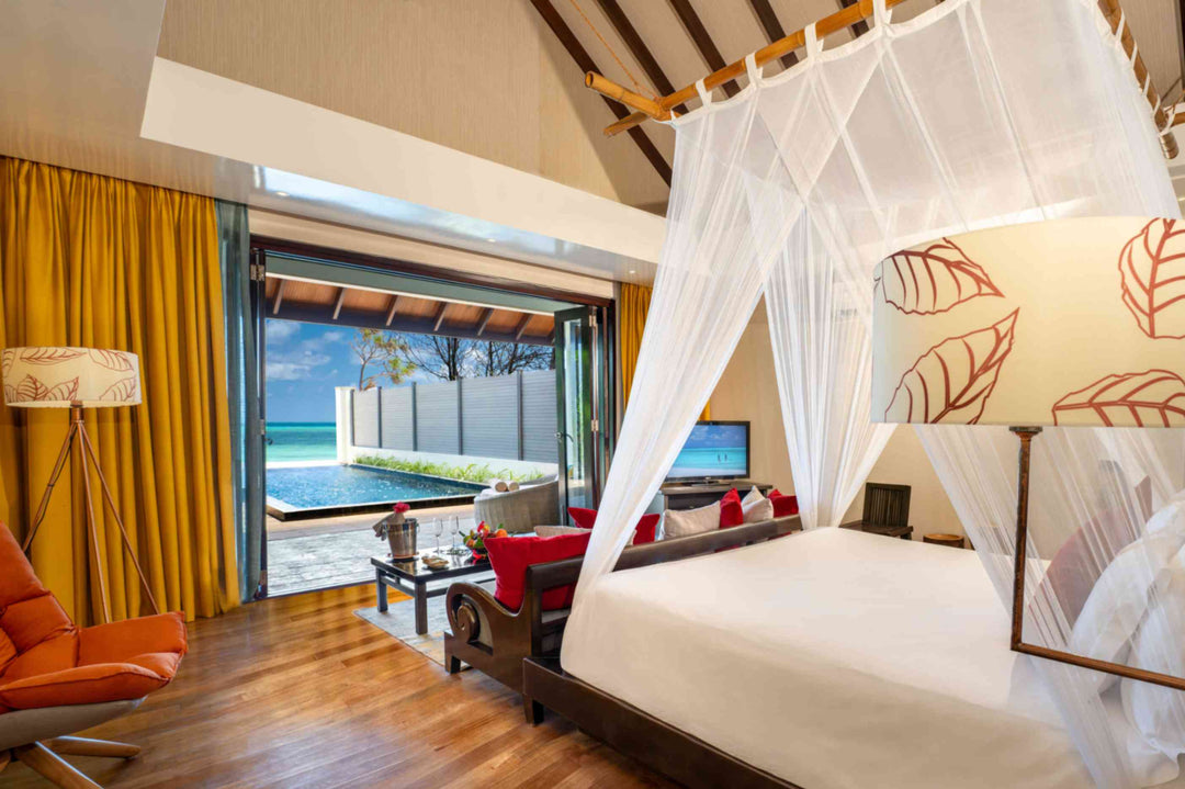 Atmosphere Kanifushi, Lhaviyani Atoll — bedroom of a Kanifushi Beach Villa with Pool: a four-poster bed under a mosquito net, red and orange cushions, doors open on to the pool.