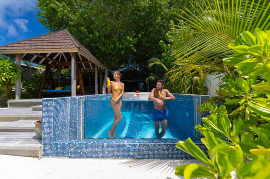 Atmosphere Kanifushi, Lhaviyani Atoll — two guests standing in the pool of a Kanifushi Grand Pool Villa beside its timber sala, surrounded by tropical planting.