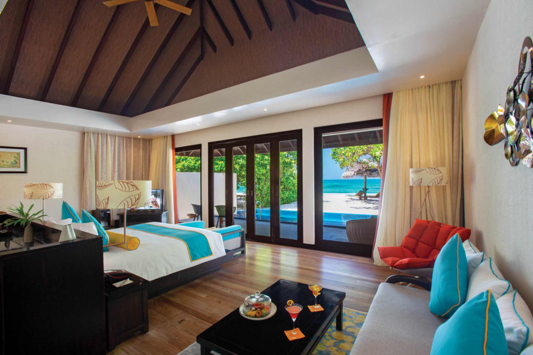 Atmosphere Kanifushi, Lhaviyani Atoll — a pool villa bedroom with turquoise and red cushions, glass doors folded open on to the pool deck and the beach.