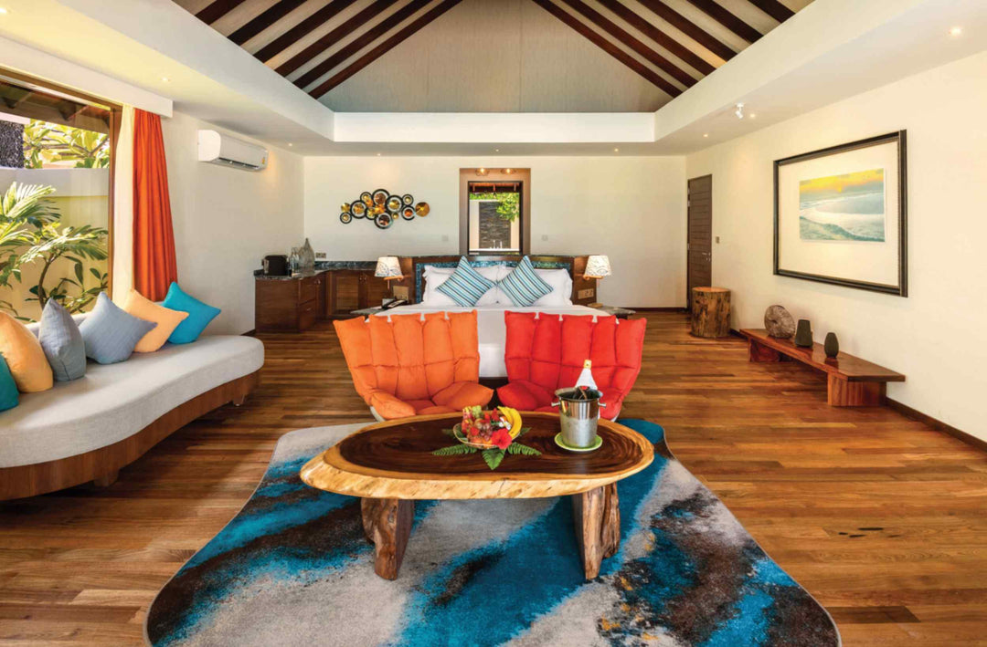Atmosphere Kanifushi, Lhaviyani Atoll — interior of a Kanifushi Grand Pool Villa: a bed with orange and blue cushions, a red armchair and a live-edge table set with fruit.
