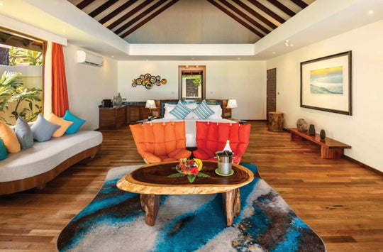 Atmosphere Kanifushi, Lhaviyani Atoll — interior of a Kanifushi Grand Pool Villa: a bed with orange and blue cushions, a red armchair and a live-edge table set with fruit.