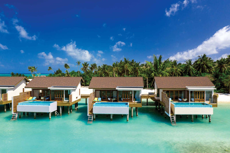 A row of three Sunset Water Villas with Pool at Atmosphere Kanifushi on stilts over a pale turquoise lagoon, with the island's palms close behind them.