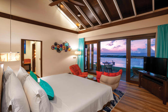 Atmosphere Kanifushi, Lhaviyani Atoll — the bed in a Sunset Water Villa with Pool facing open doors on to the deck and the ocean at sunset.