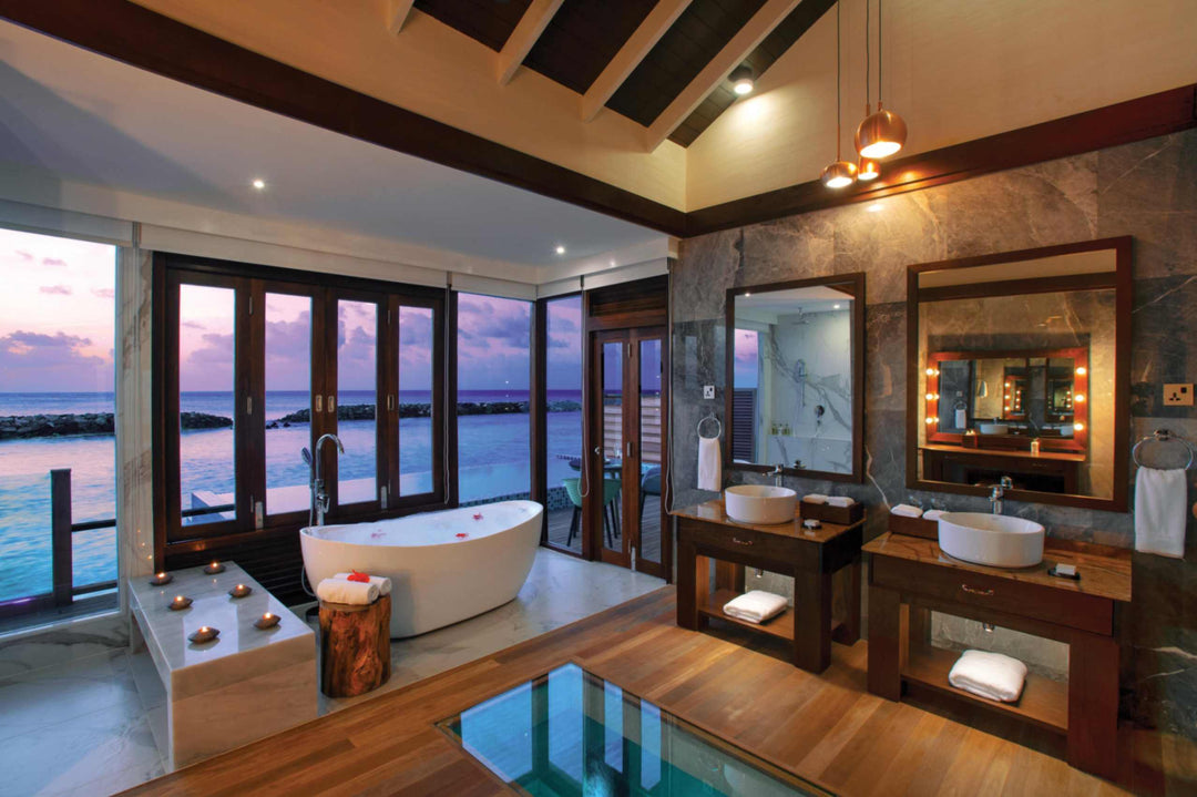 Atmosphere Kanifushi, Lhaviyani Atoll — bathroom of a Sunset Water Villa with Pool: a freestanding bath and twin stone basins in front of windows on to the ocean at dusk.
