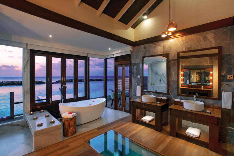 The bathroom of a Sunset Water Villa with Pool at Atmosphere Kanifushi, a freestanding bath and twin stone basins in front of windows on to the ocean at dusk.