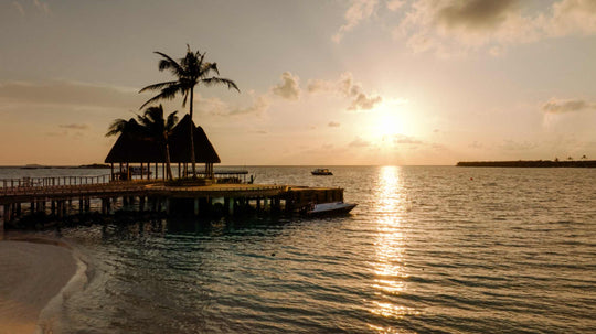 The sun setting beyond the overwater restaurant at ananea Madivaru Maldives, seen from the beach