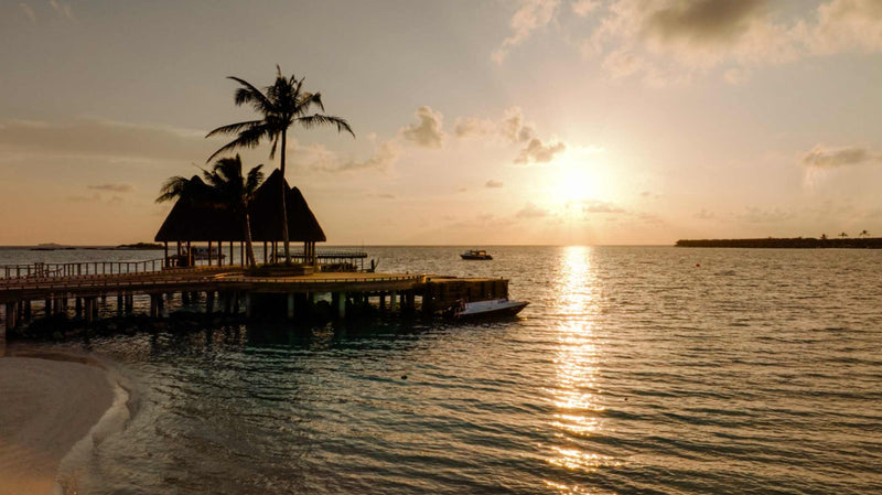 The sun setting beyond the overwater restaurant at ananea Madivaru Maldives, seen from the beach