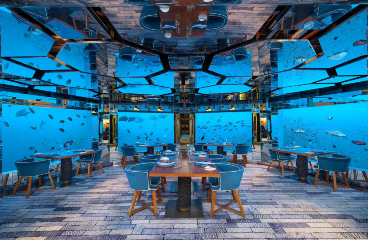 The dining room of SEA at Anantara Kihavah Maldives Villas, tables set beneath glass walls full of reef fish and a mirrored ceiling