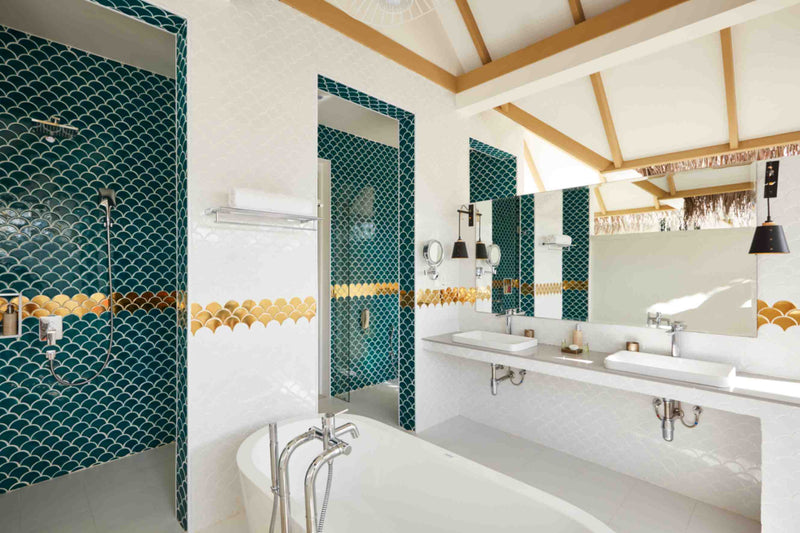 A bathroom at Sun Siyam Iru Veli in teal fish-scale tiling with a walk-in shower, twin basins and a tub under a thatched ceiling.
