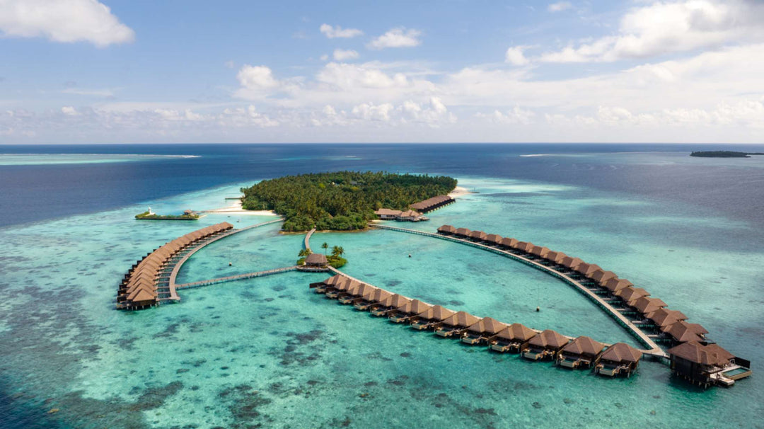 Ayada Maldives from the air, the wooded island on one side and both arms of overwater villas curving into the lagoon