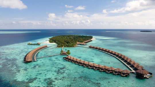 Ayada Maldives from the air, the wooded island on one side and both arms of overwater villas curving into the lagoon