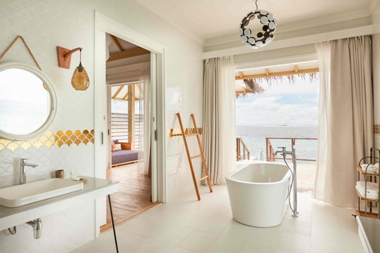 Sun Siyam Iru Veli, Dhaalu Atoll — bathroom with a round mirror and gold tile band, tub, and the bedroom and lagoon beyond