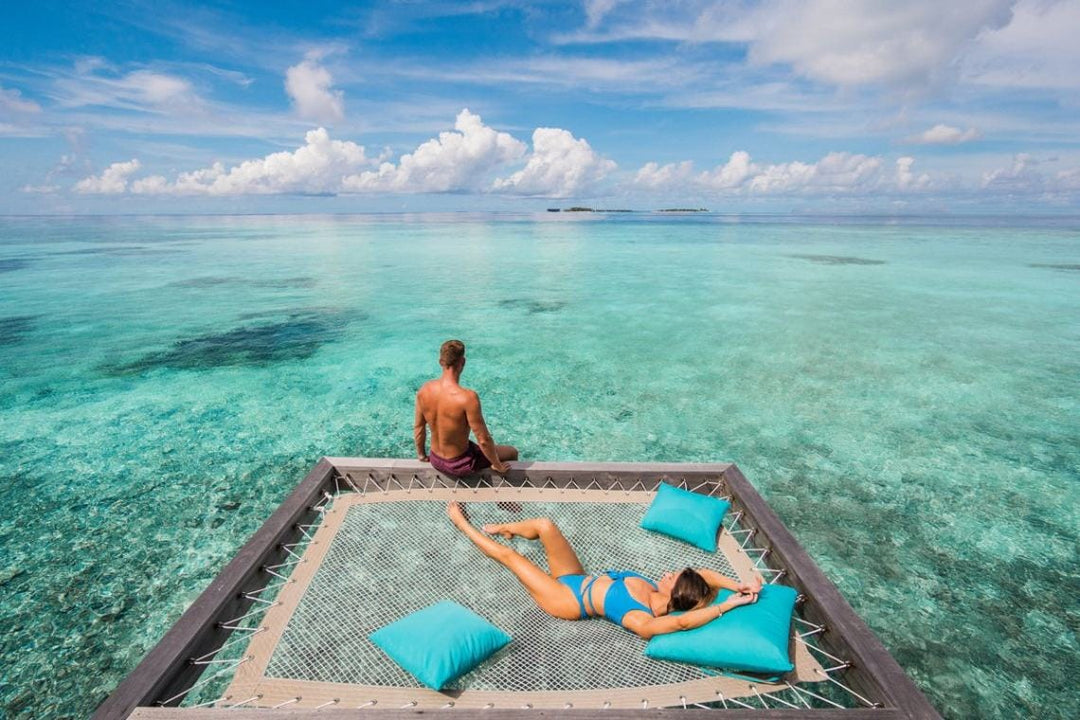Hurawalhi Maldives, Lhaviyani Atoll — a couple on the net hammock of an overwater villa, clear reef water below and an island on the horizon