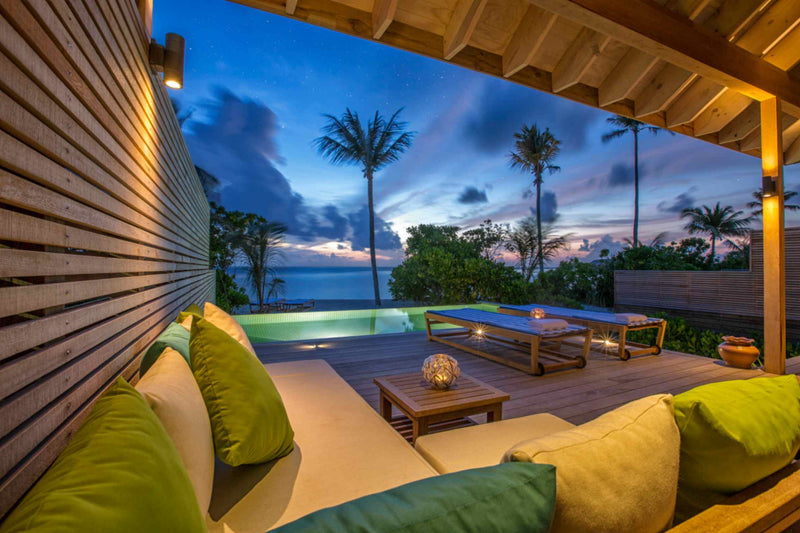 A villa terrace at Hurawalhi Maldives at dusk, lanterns lit on the deck and the pool glowing green against a violet sky.
