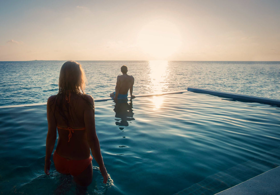 Hurawalhi Maldives, Lhaviyani Atoll — two guests at the infinity edge of a pool at sunset, the sun low over open sea beyond