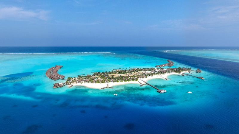 Aerial view of JW Marriott Maldives Kaafu Atoll Island Resort, the wooded island ringed by a pale lagoon with overwater villas along one side